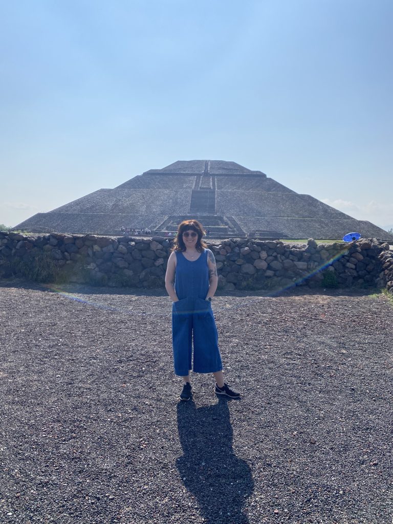 Siân Evans wearing a blue jumpsuit and sunglasses, standing in front of one of the pyramids at Teotihuacan.