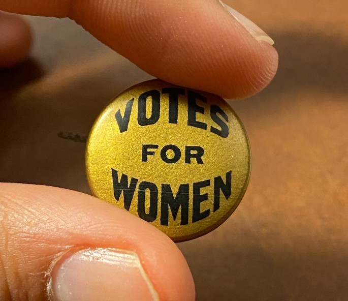 Fingers holding a small pin with the slogan "votes for women" printing on it.