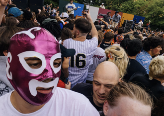 photograph of a crowd in front of a band playing live music, with the person in the foreground wearing a purple and white luchador mask