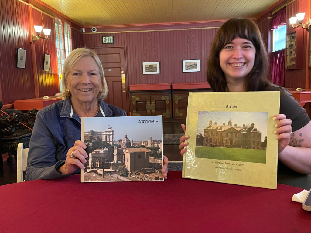 Mary Meyer and Michelle Fitzgerald sit at a table in Evergreen's Far East Room. Each is smiling at the camera while holding a photo album from Mary Meyer's Attingham experiences. 