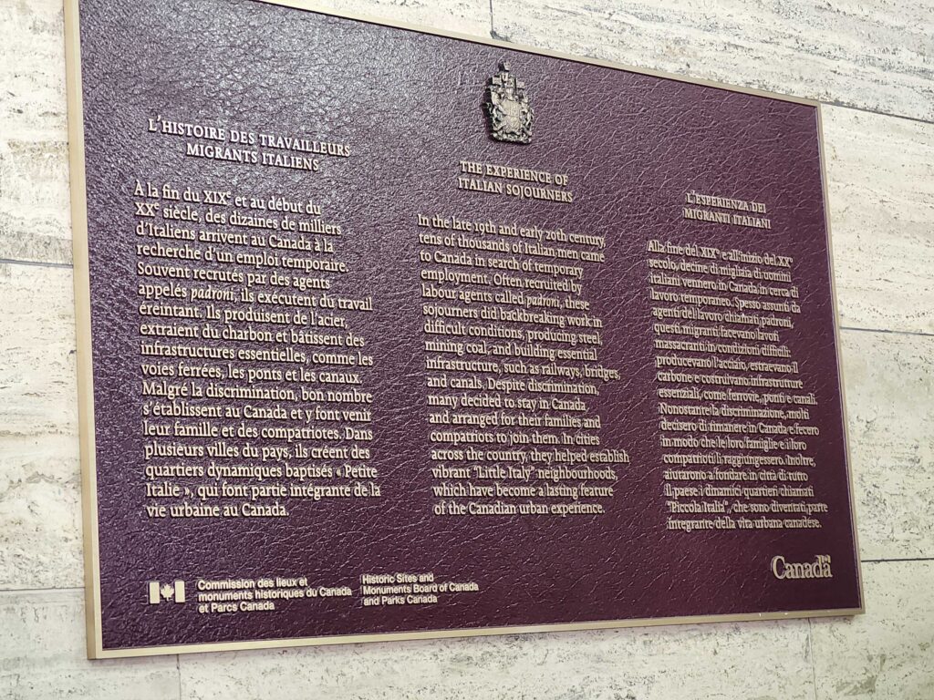A burgundy plaque mounted to a stone wall. In three columns, it explains the story of the experience of Italian sojourners. The first row is in French, the second row in English, and the last row in Italian. 
