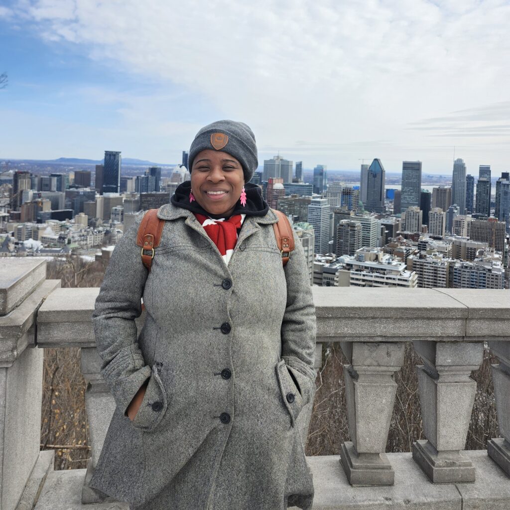 A young black woman in her 30s stands in front of a city skylight with a stone railing behind her in late winter/early spring. She wears a long grey coat, a grey cap, and a red and white striped scarf. She smiles at the camera. 