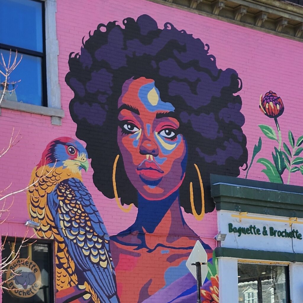 A portrait of a dark skinned woman with a fro with a falcon sitting in her hand. It has been painted as a mural on the side of a building using vibrant purples, blues, red, and gold colors. 